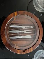 Top view of round wooden cheese board with glass cover showing cheese utensils inside (two knives and one fork)