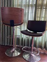 Photo showing the set of two adjustable MCM bar stools, one with the front seat and black cushion visible, the other showing the wood veneer back, both with chrome pedestal bases and footrests.