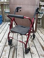 Front left perspective of a red rolling walker with padded seat and hand brakes, showing both front and rear wheels and seat back support.