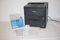 Front view of the Brother HL-5470DW printer next to the Brother TN-750 toner cartridge box and test print pages.
