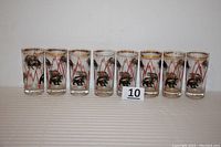 Photo showing all eight clear glass highball tumblers in a row with black and gold bull and bear decorations and red stock market graph lines, gold rims with visible wear.