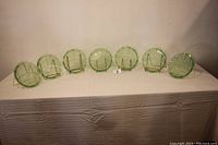Six green Depression glass three-part grill plates arranged side by side, showing scalloped edges and three divisions on each plate