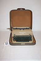 Beige Remington Rand typewriter inside an open hard case with handle, resting on a striped surface, front view showing brand name and keyboard.