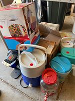 Assorted plastic pitchers, insulated dispenser, storage containers and jars in various colors including blue, white, red, and turquoise, arranged together with box visible in background.