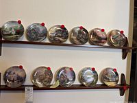 Photo showing 12 Thomas Kinkade collectible plates arranged on two wooden wall shelves.