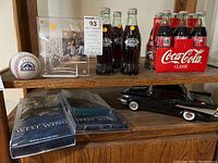 Wide view of shelf showing Colorado Rockies baseball collectibles including signed baseball, acrylic figurine case, Coca-Cola bottles in cardboard carriers, black model car and West Wing DVD box sets.