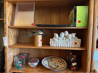 Wooden shelf holding various decor items including figurines of birds and a group of people, a woven bowl, butterfly lamp, and glass containers with red stickers.