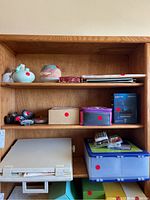 Wooden shelf with various decorative pottery items on top shelf, small boxes, electronic items in box, plastic storage containers, and an office ledger binder on lower shelves.
