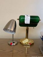 Two small desk lamps side by side on a wooden surface. Left lamp is small metal with conical shade, right lamp has green duckbill glass shade on gold-tone base.