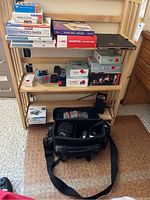 Photo of wooden shelf with stacked photo paper boxes, Canon camera boxes, camera bag with contents, and assorted camera accessories and batteries on shelves below.