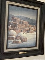 Full view of framed painting showing Pueblo architecture in snow under a dark wooden frame with a beige inner matting.