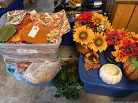 Three clear storage bins filled with fall-themed table linens and various decor items, a few artificial flower arrangements in pumpkin containers, and small decorative figurines arranged on top and nearby, along with some artificial greenery scattered on the floor.