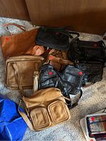 10 mostly leather purses and bags arranged on carpet; various styles and colors including black, tan, and cream; some have external zippered compartments and different strap types