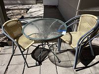 Photo showing two rattan-backed chairs positioned around a round glass-top patio table with a metal frame on a paved outdoor surface.