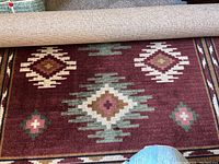 Photo showing rolled and unrolled rug with southwest geometric diamond pattern in maroon, green, cream, pink, and brown colors.
