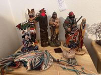 Overview shot of the 8 assorted figurines and textile art pieces displayed on a table including totem poles, frontier soldier figures, a painted pottery figurine, and textile craft items.