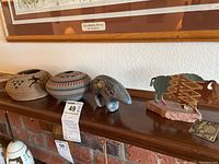 Five southwestern-style decor pieces displayed on wooden mantle: two pottery vessels, grey ceramic animal sculpture, metal buffalo figurine on stone base, and cut rock lidded box.