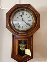 Close-up photo of wooden regulator wall clock showing dial with Roman numerals and pendulum behind glass door.