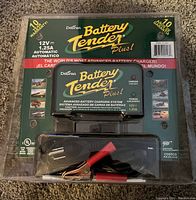 Front view of packaging showing Deltran Battery Tender Plus charger with cables inside transparent blister pack, highlighting specifications and warranty.