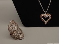 Photo showing the rose-gold-plated heart pendant necklace on a black display with the arabesque ring positioned to the side showing overall lot items