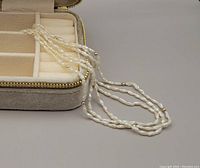 Three-strand rice pearl necklace partially inside the velvet storage/travel box with divided interior compartments.