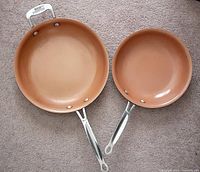 Top down view of both red copper frying pans laid on carpet showing interior surfaces and handles.