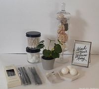 Overview of the items included in the bathroom decor lot showing the apothecary jar with decorative filler, small jars with cotton balls and Q-tips, faux plant, framed sign, tealight candles, incense sticks, night light, and bath bomb tray.