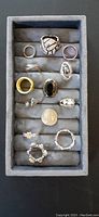 Top down view of 13 rings arranged in a grey jewelry display tray, showing variety in size and style.