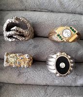 Four men's fashion rings displayed, showing varied designs and materials.