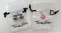 Two Thomas Sabo charms on white drawstring bags with logo, showing Love Bug van charm with a peace sign and 'LOVE' text, and a pink peace sign charm.