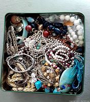 Top view of a metal box filled with assorted costume jewelry pieces including chains, beads, and pendants in various colors and materials.