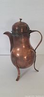Side view of vintage copper kettle showing curved body, handle, hinged lid with knob, and one leg