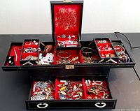 Full view of black jewelry box open showing red velvet compartments filled with assorted vintage jewelry including pins, cufflinks, bracelets, earrings