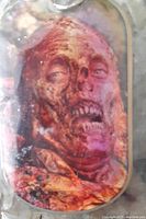 Close-up of a Walking Dead zombie face dog tag showing detailed monstrous features, partly decomposed with teeth exposed.