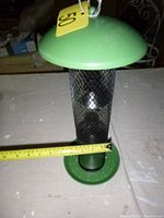 Side view of metal tubular bird feeder with mesh body and green metal top and base, with measuring tape showing size.