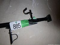 Photo of black adjustable metal curtain rod with green lot number tape. Shows mounting bracket and square finial end, length adjustment mechanism visible.