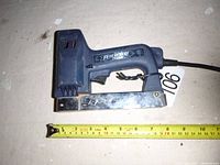 Side view of Arrow Electric stapler ETF50BN on a surface with a yellow measuring tape showing length approx. 7 inches.