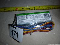Photo of electronic ballast showing front label with specifications and wiring bundled, placed next to a measuring tape for size reference.