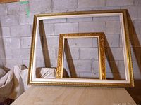 Two wood frames leaning against a concrete block wall, the larger frame is white and gold and the smaller frame is fully gold-toned with carved designs.