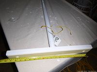 White metal adjustable support pole lying horizontally on a surface with a measuring tape showing the width of the wide rectangular base around 19 inches.