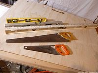 Three levels and two hand saws arranged on a plywood surface. Two hand saws show wear, one large saw is warped.