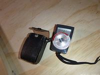 Sekonic Auto-Lumi Model 86 light meter device featuring front dial and attached wrist strap, with black leather carrying case beside it on a wood surface.