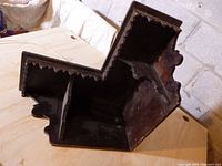 Side view of the old wooden corner shelf showing the carved decorative edges and worn surface, placed on a wooden table against a block wall.