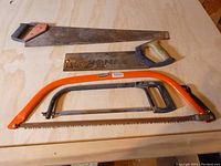 Four old saws laid out on wooden surface: 26 inch hand saw, 13 inch hand saw, CutBrik Portugal swede saw, orange framed hacksaw