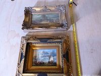 Two gilt-framed paintings stacked with measuring tape visible