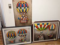 Three framed paintings resting against a wall. Each artwork features vibrant, colorful hot air balloons with cartoonish human figures in the baskets playing musical instruments.