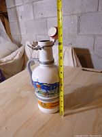Side view showing stein height next to measuring tape at about 12 inches tall, with colorful winter Christmas village scene on white ceramic body, metal handle and lid clasps.