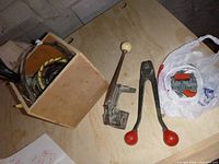 Photo showing metal strapping tensioning and sealer tools, wooden box containing metal connectors and scrap strapping on wooden surface.
