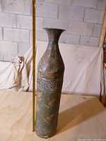 Full view of the thin metal vase measuring about 26 inches tall with a flared top and decorative pierced metal bands on a mottled patina surface.