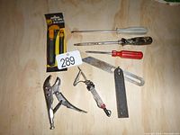 Photo showing seven small hand tools arranged on plywood including three screwdrivers, locking pliers, a metal square, a utility knife in package, and a test light screwdriver.